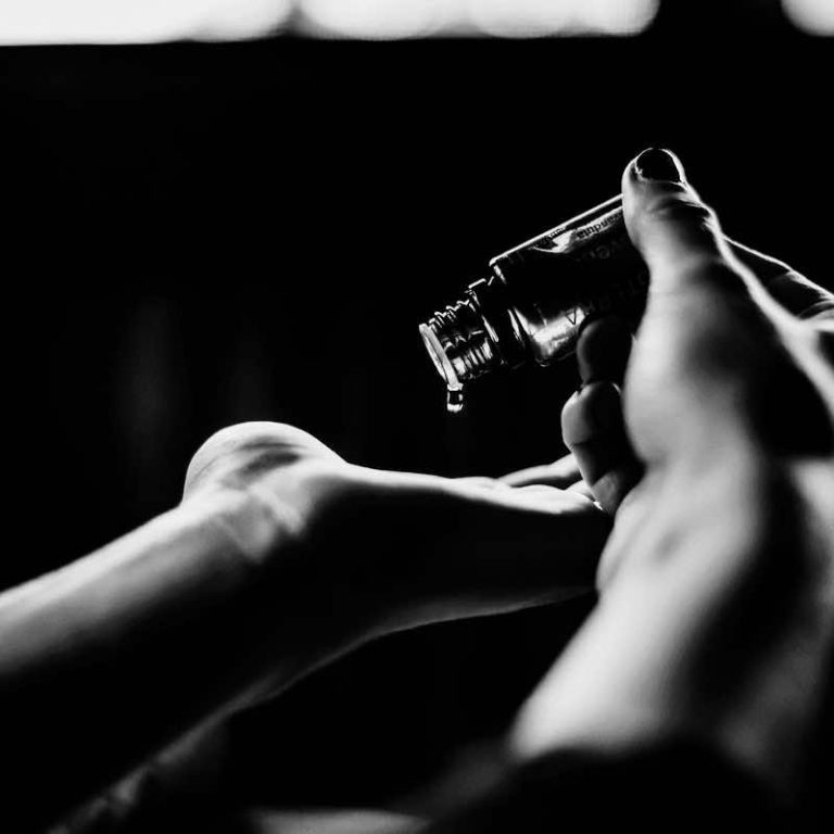 A person holding a small bottle, pouring liquid into their other hand in soft light.