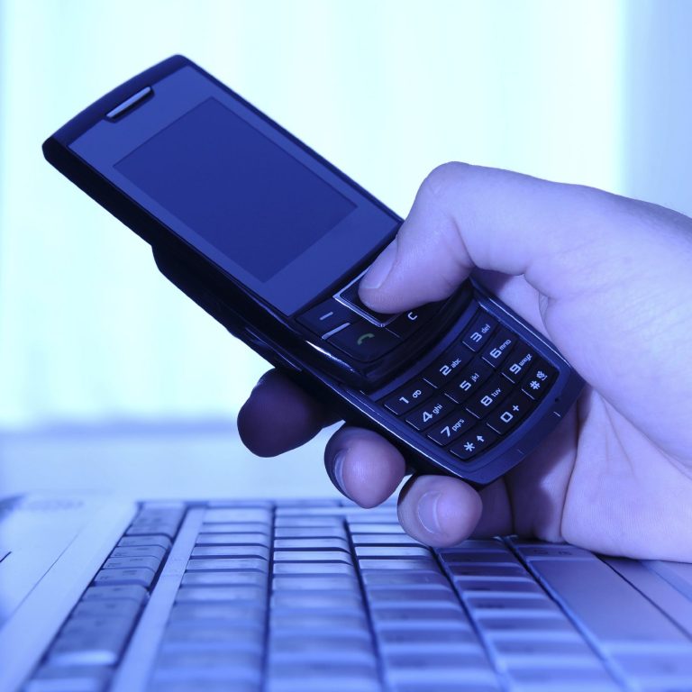 A hand holding a black mobile phone above a laptop keyboard.
