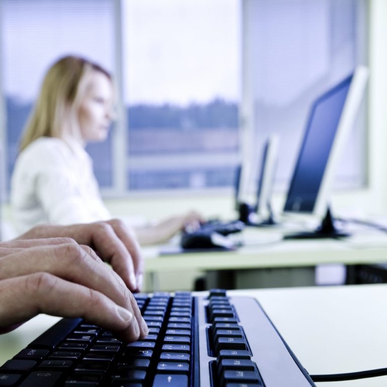 A woman types on a keyboard with a computer in the foreground and another in the background.